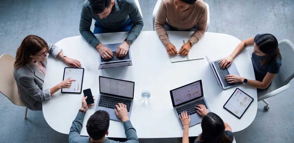 A diverse team of professionals around a conference table using multiple digital devices to collaboratively review and analyze real-time work performance data.